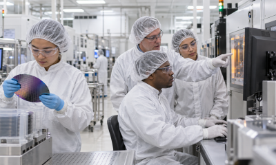 A semiconductor instructor guides three technicians as they inspect a silicon wafer and review manufacturing equipment inside a cleanroom training facility.